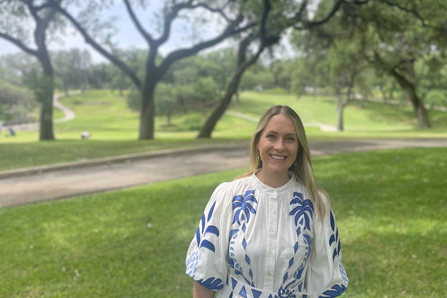 A blond woman in a white and blue dress stands outside in front of oak trees.