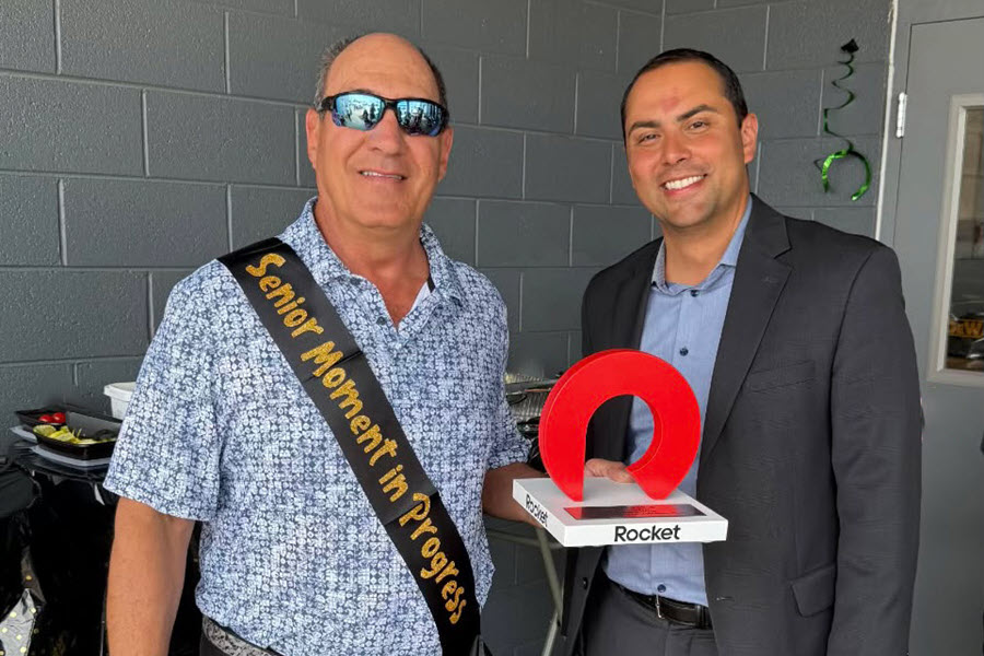 Two men stand next to each other. One is wearing a retirement sash and the other is holding a golf trophy.