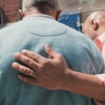 The back of two men. One has his hand on the other's back.